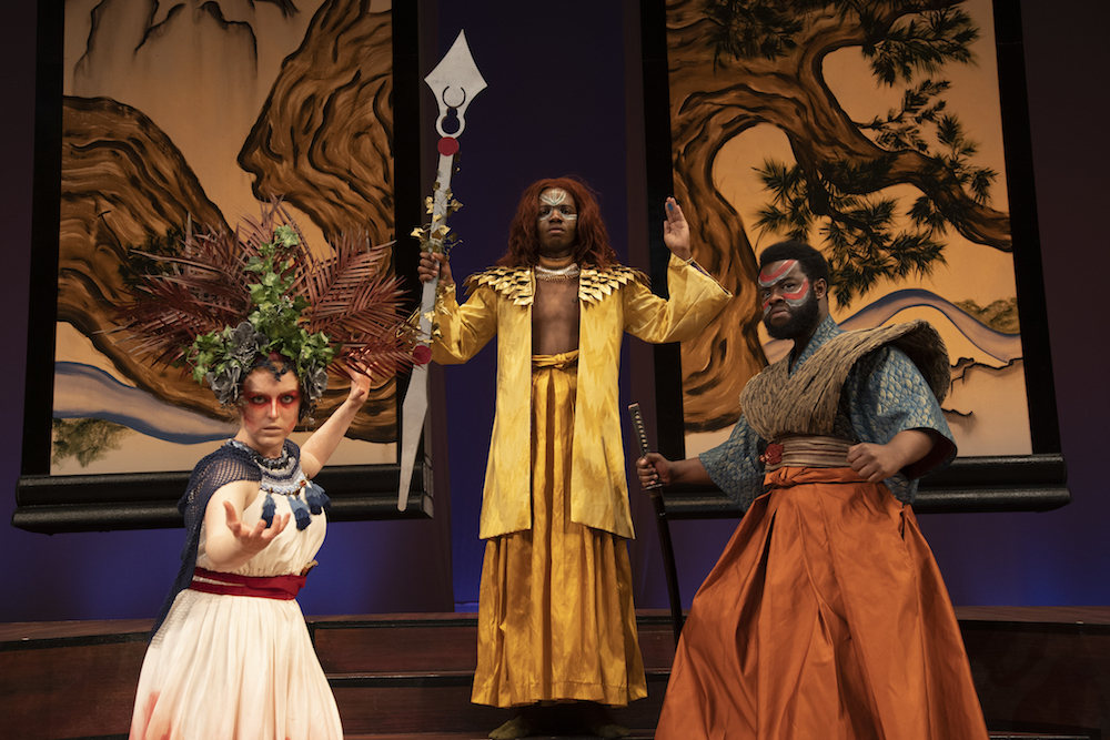 Three actors posing in costume. Actor on the left is wearing a leaf headress and a cream dress. Dionysus is center adorned in gold and wielding a staff. The right individual is wearing red face paint, brown chest-plate and shoulder pads with an orange skirt, and is holding a sword.
