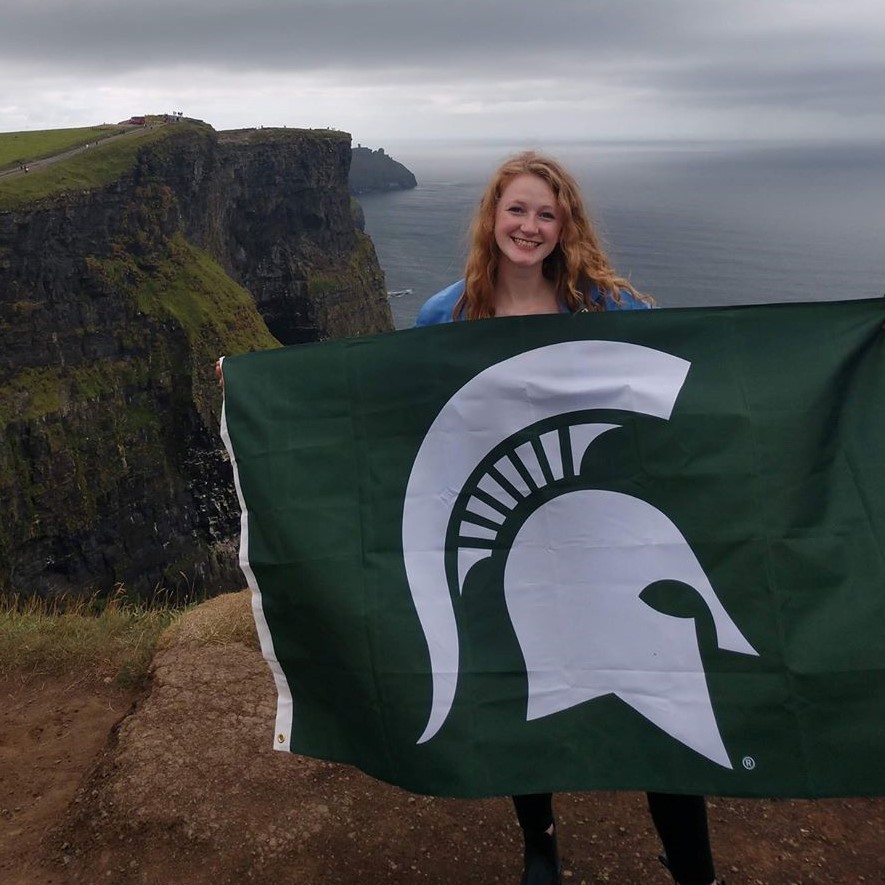 woman holding an MSU flag standing in front of a cliff looking to the ocean